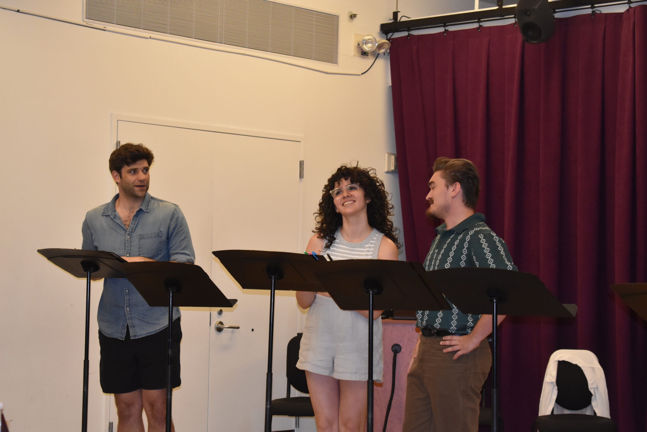 Three actors are engaged in a rehearsal, using music stands for scripts.